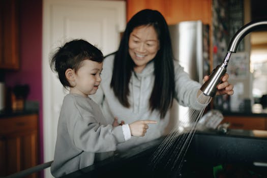 A mother and child happily wash hands together at a kitchen sink, sharing a bonding moment indoors.